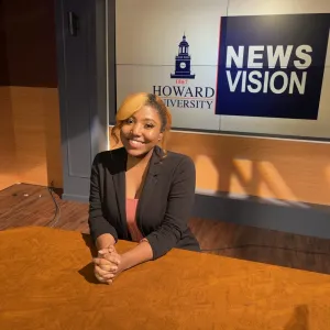 Ianna Fenton, a young woman with blonde hair, wearing a black blazer, smiles while seated at a news anchor desk with a "Howard University Newsvision" logo in the background.